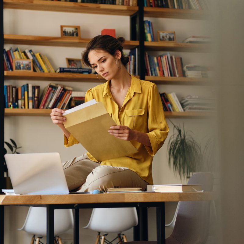Beautiful woman in shirt sitting on desk with documents thoughtfully working in modern office